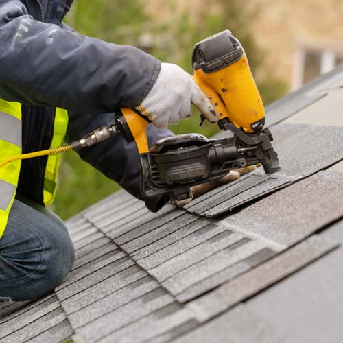Roofer in safety vest using pneumatic nail gun to install shingles
